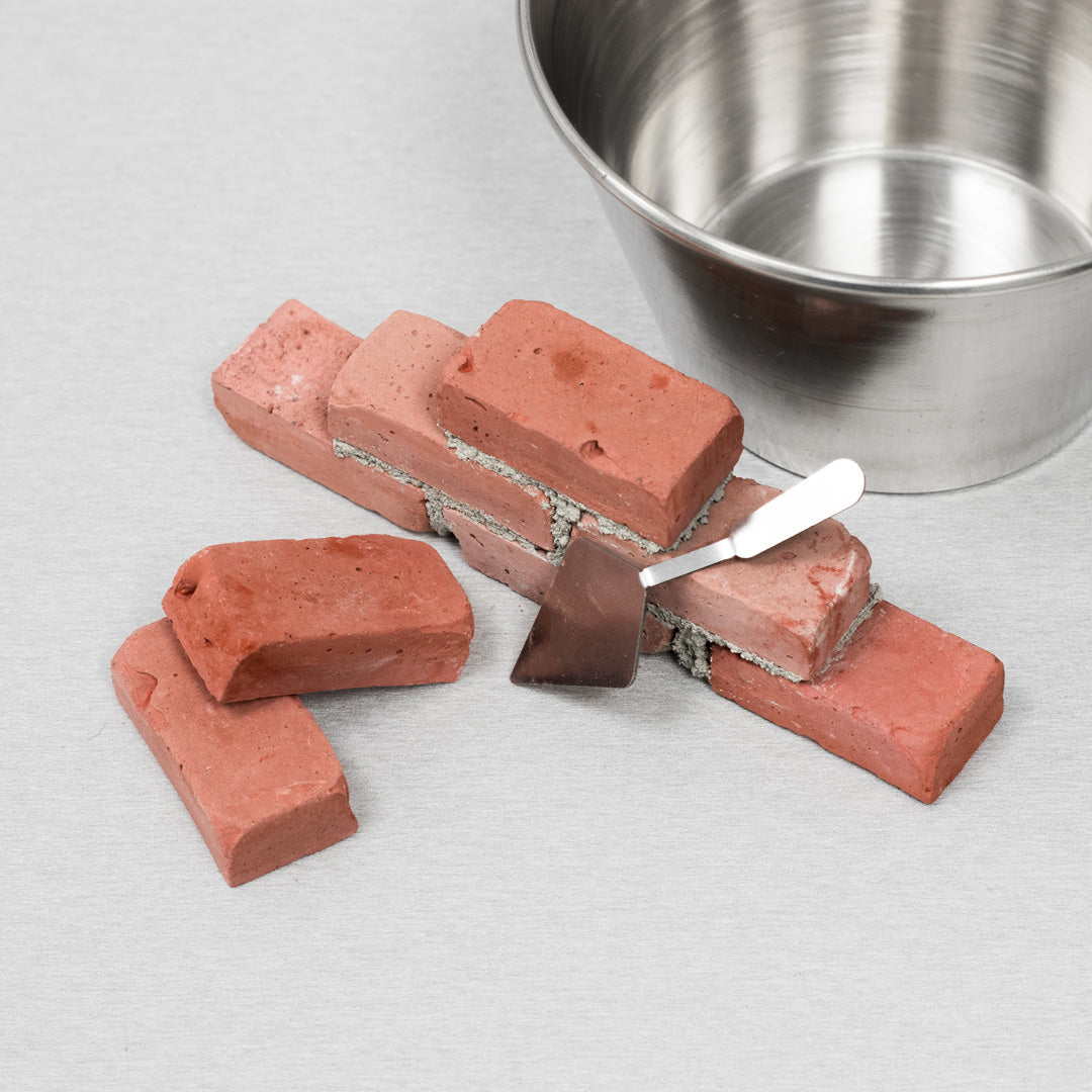 A miniature trowel placed againg small brick wall made of vibrant red, handmade mini bricks with. Background is light gray wood. Trowel is made of thin sheet metal with dimond shaped blade. Side top view. Two min bricks are placed randomly aside and a small metal bowl is behind to showcase all accesories.
